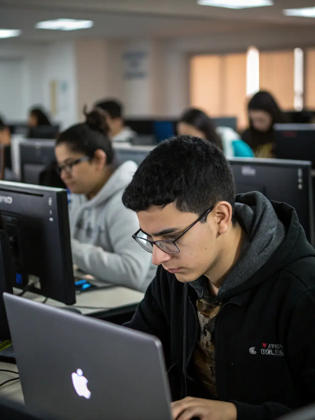 A student intently focused on a laptop screen, coding in a modern, well-lit classroom at CodeBridge Academy, surrounded by other engaged students.